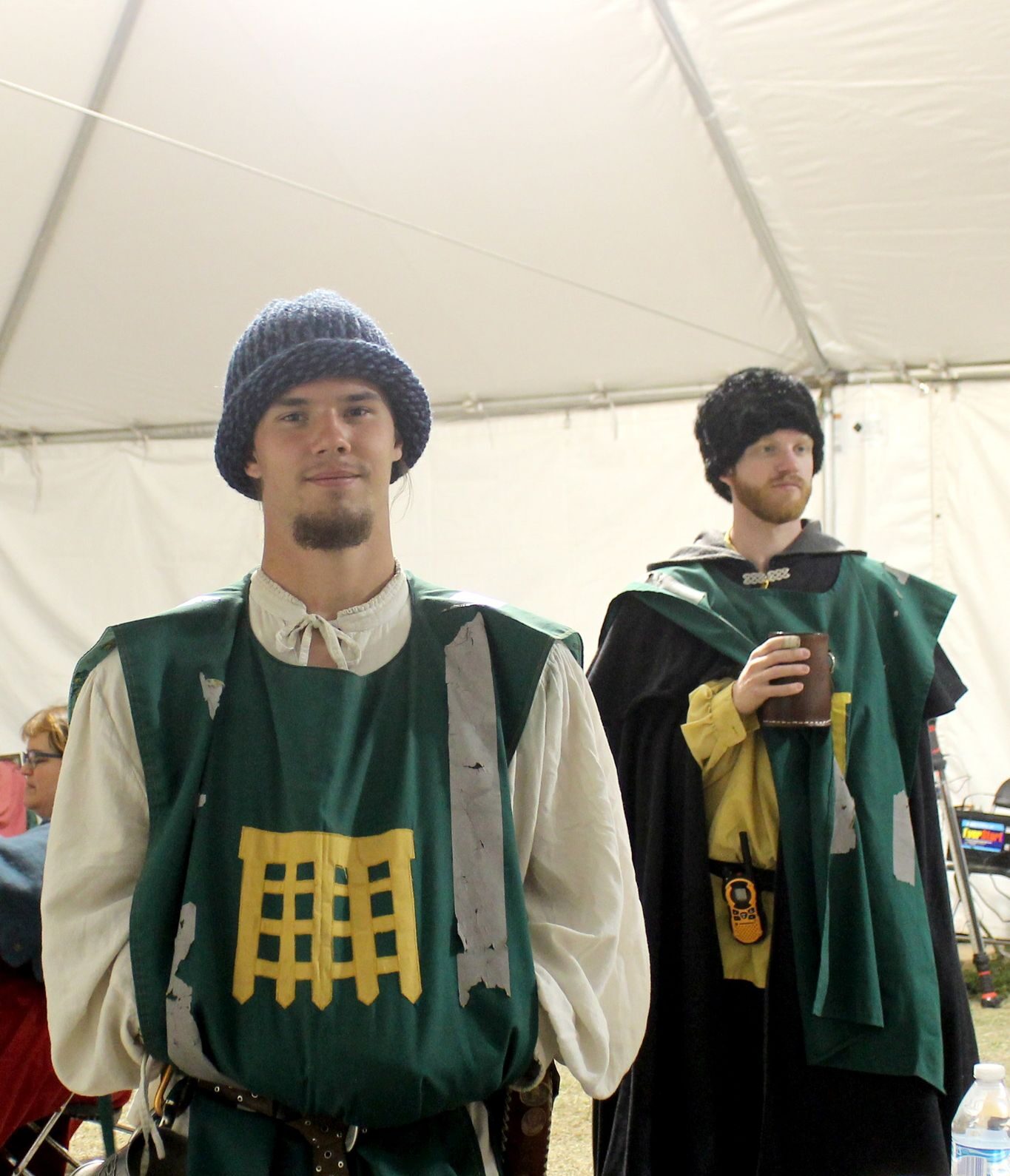 Submission 1 Two younger male presenting populace members wearing green tabbards with The Watch heraldry embroidered and displayed in gold above their attire. Both wearing cold weather tunics and caps.